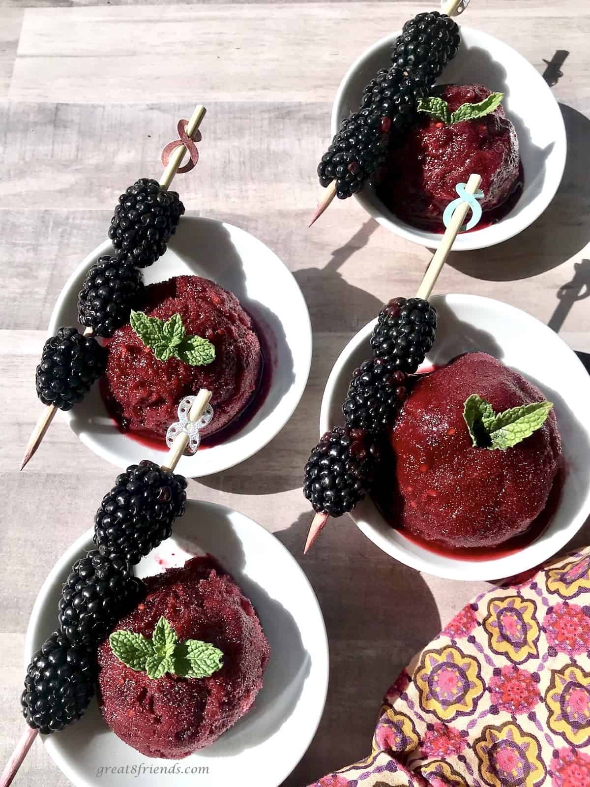 Four scoops of Blackberry Italian ice in four white bowls with fresh blackberries on sticks on the side of each bowl.