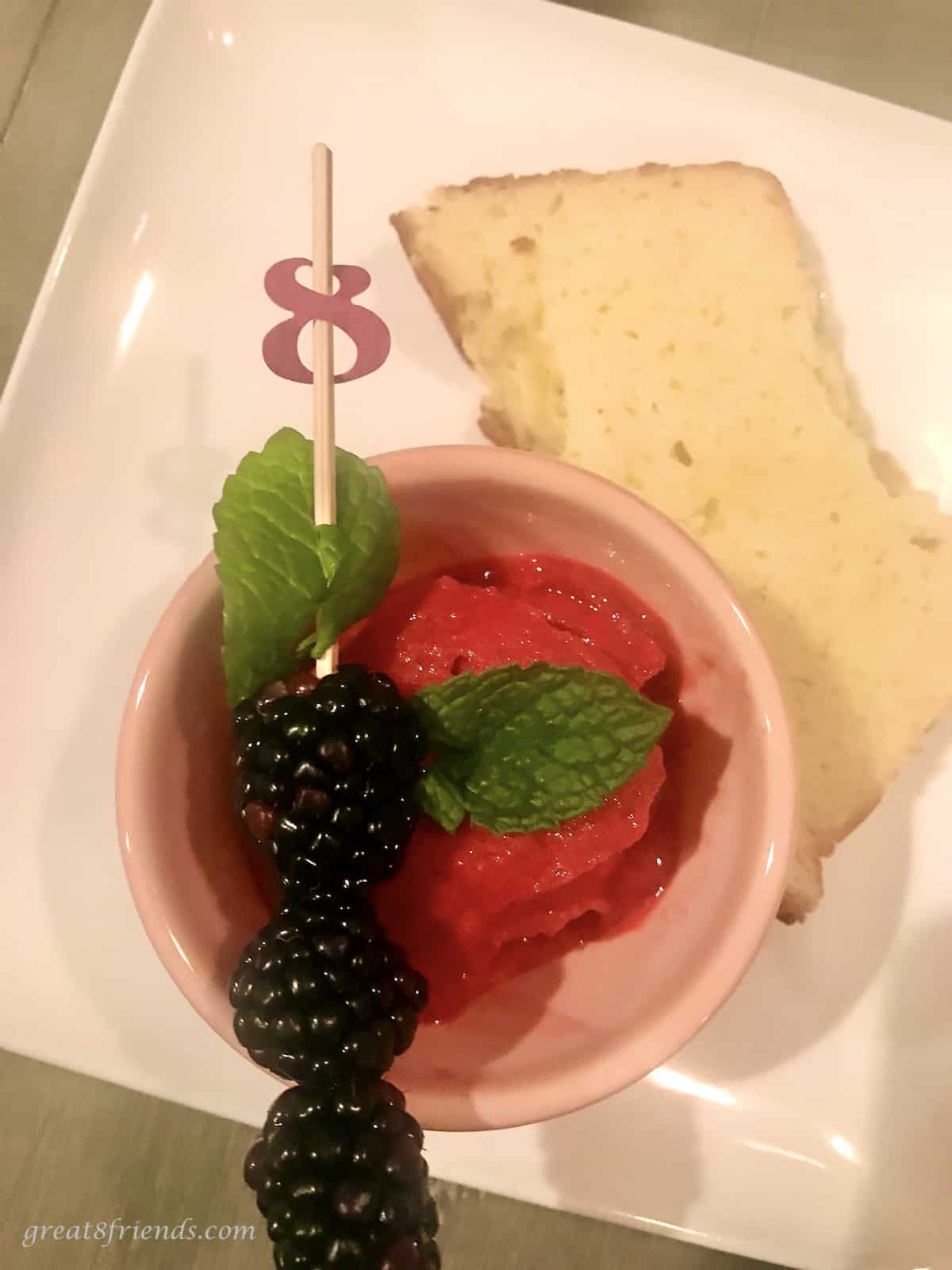 White plate with a slice of pound cake and a small bowl of blackberry ice with fresh blackberries on a stick on the edge of the bowl.