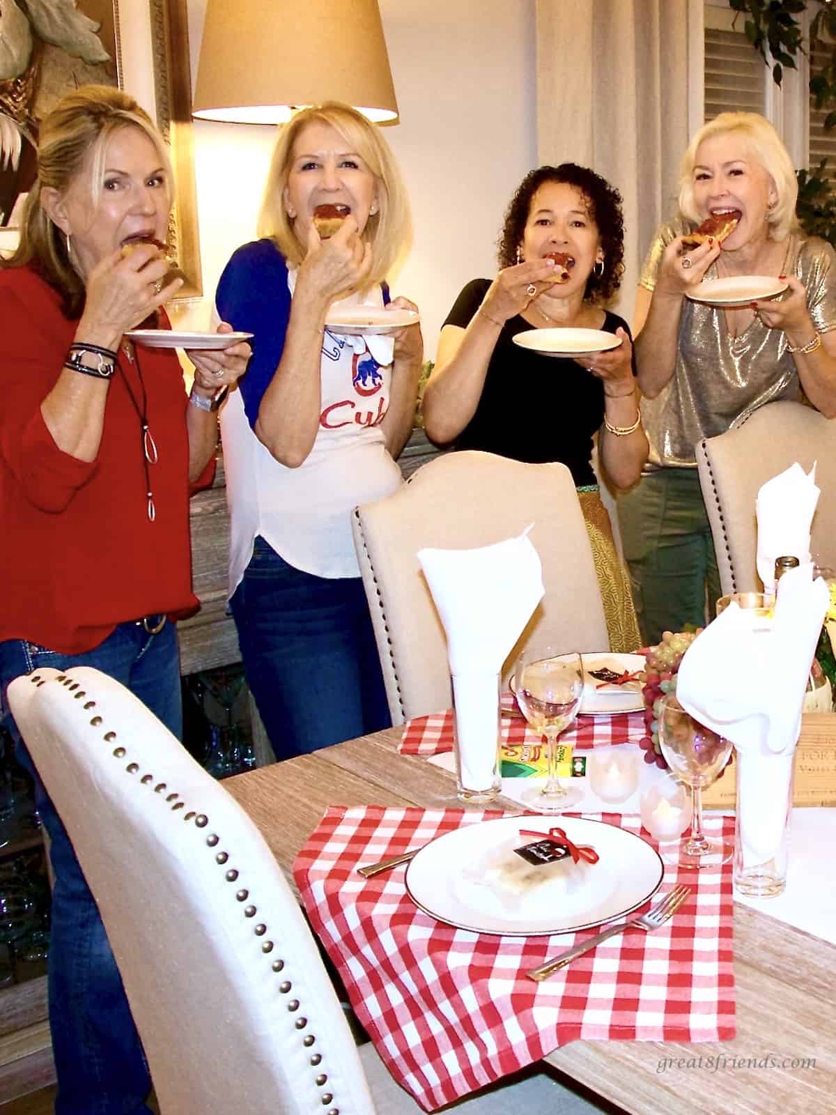 Four women eating each eating a piece of pizza standing in a dining room with a table set for an Italian meal.