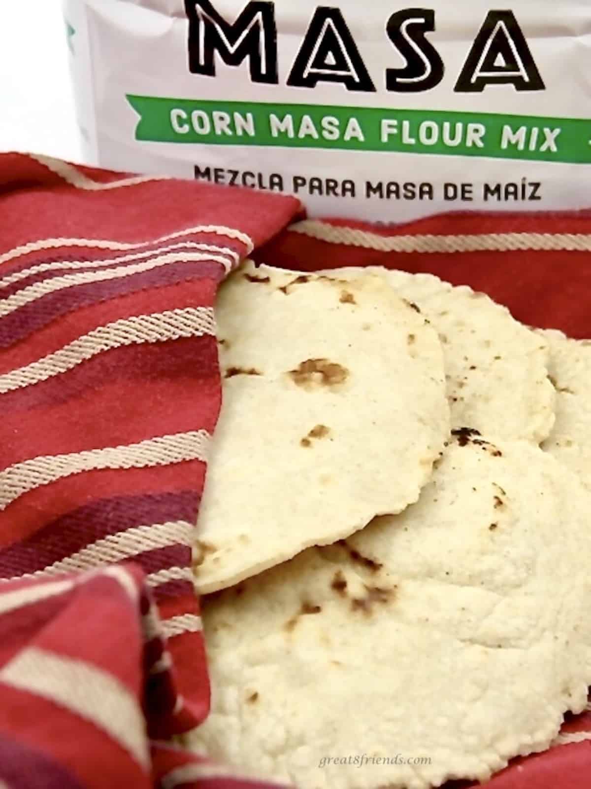 A red towel holding several corn handmade tortillas with a bag of masa flour mix in the background.