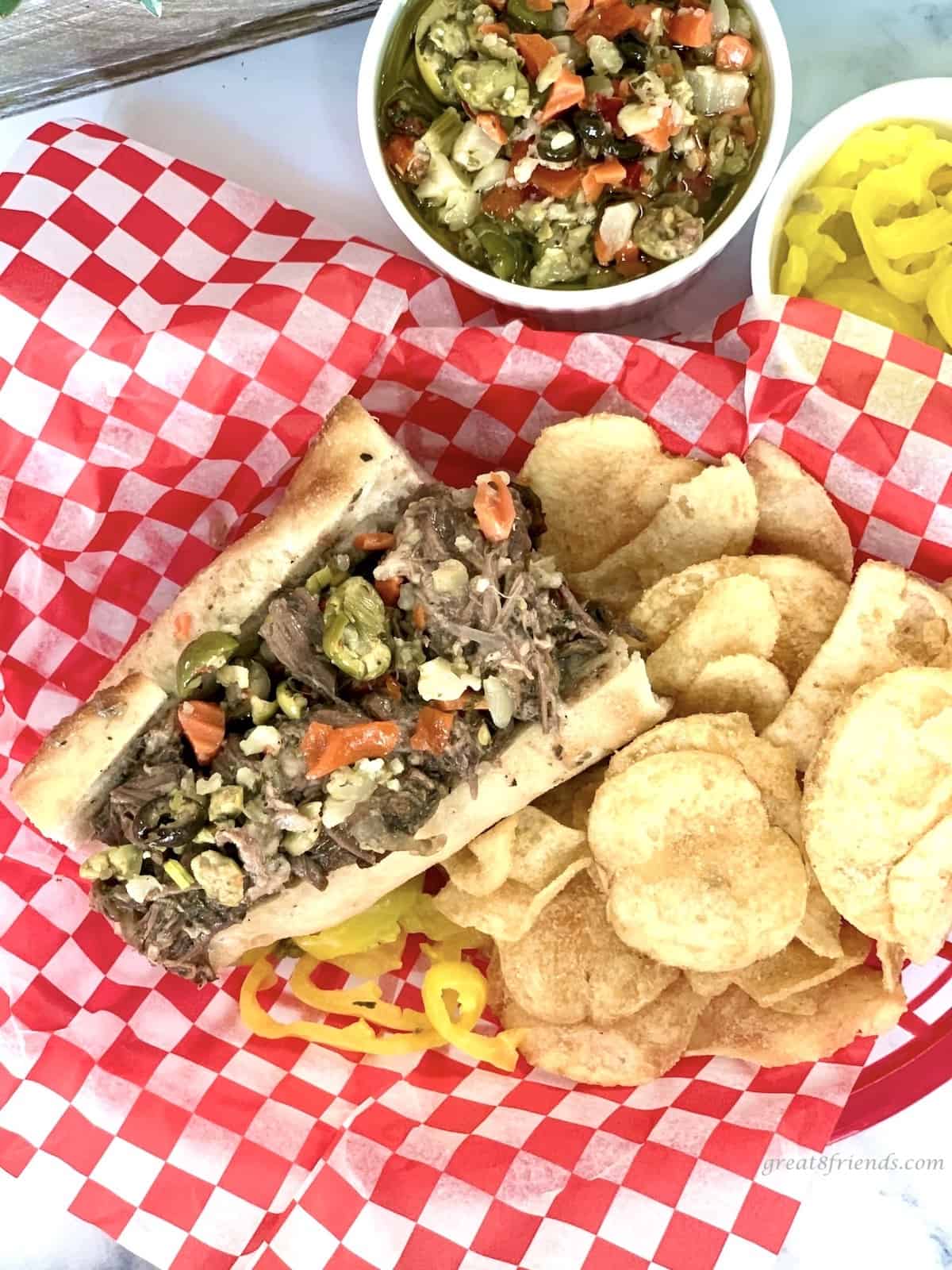 Italian Beef Sandwich in a basket with a red and white paper liner and potato chips, peppers and Giardiniera on the side