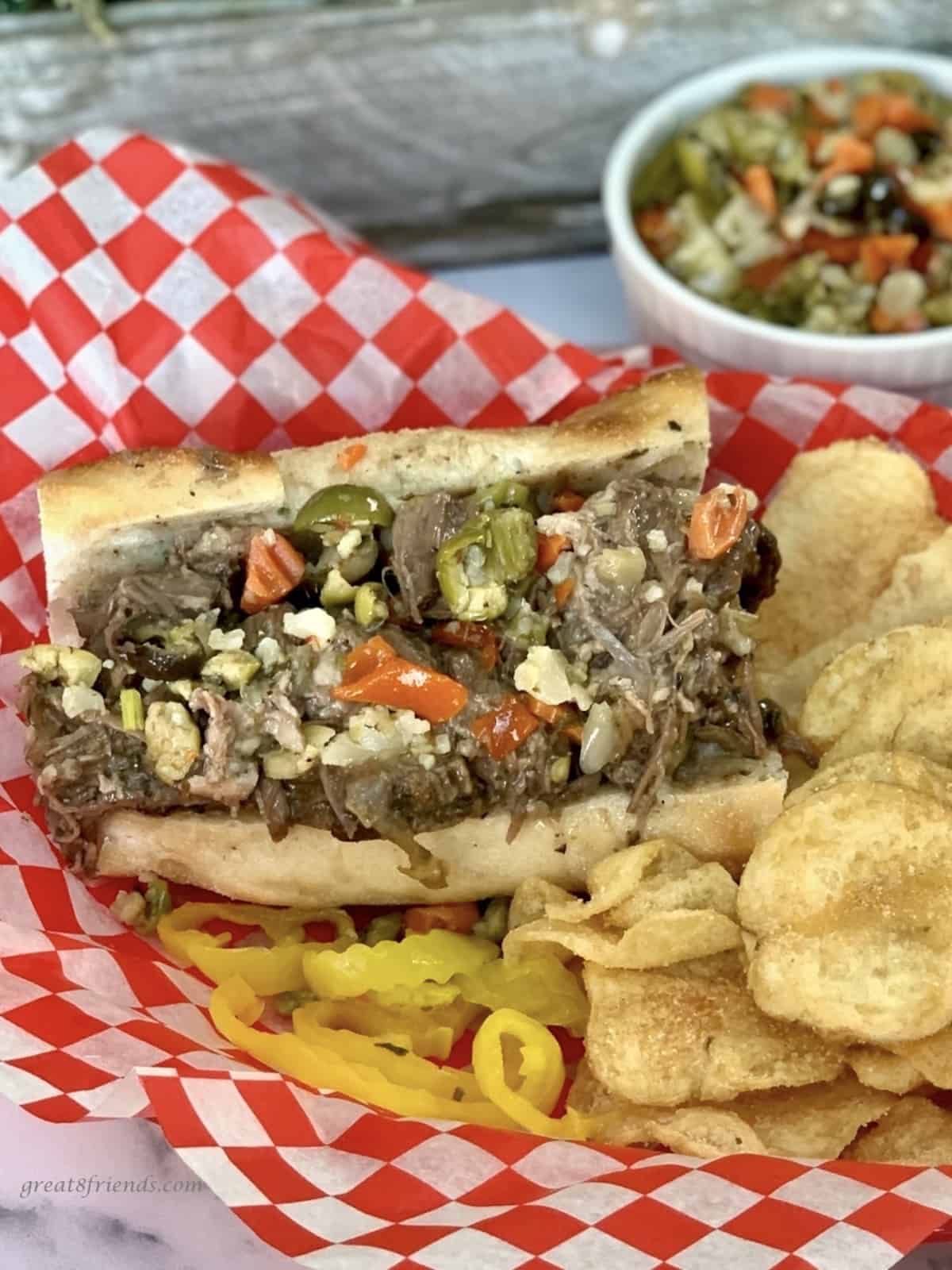 Chicago Italian Beef Sandwich in a basket with a red and white paper liner and potato chips, peppers and Giardiniera on the side.