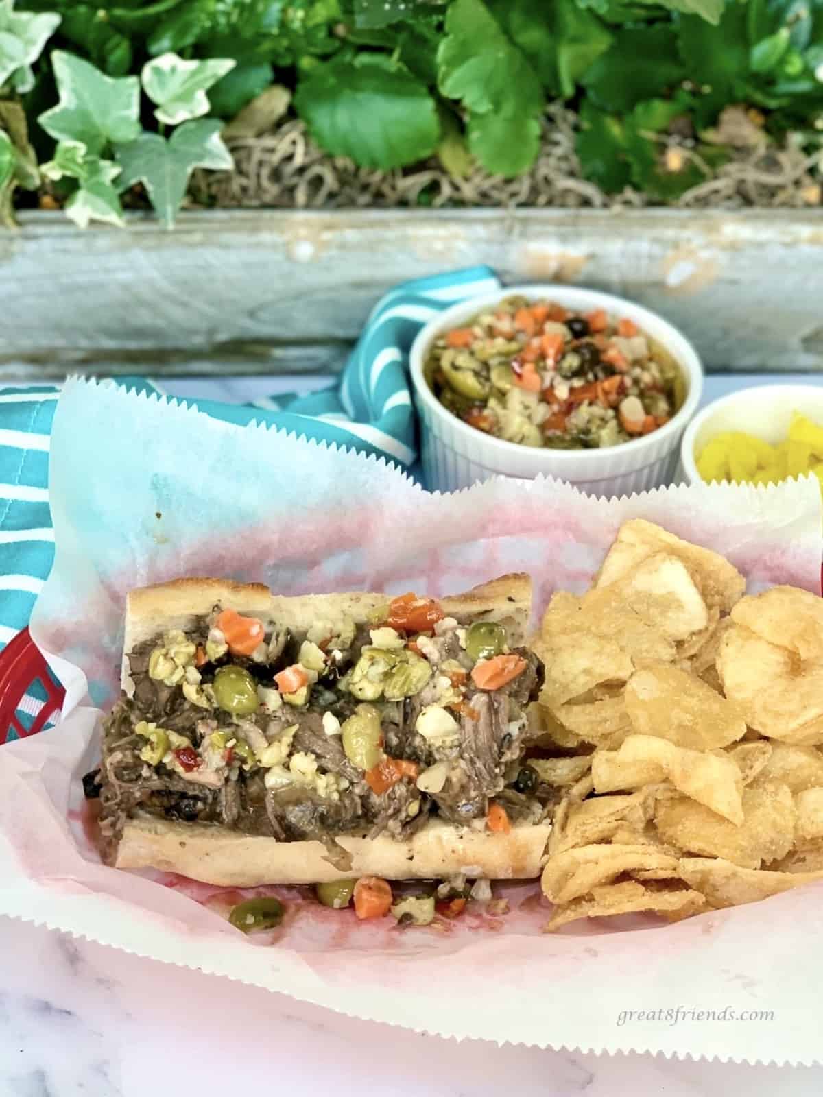 Slow cooker Italian beef sandwich with potato chips on the side and Giardiniera and deli-sliced pepper rings.