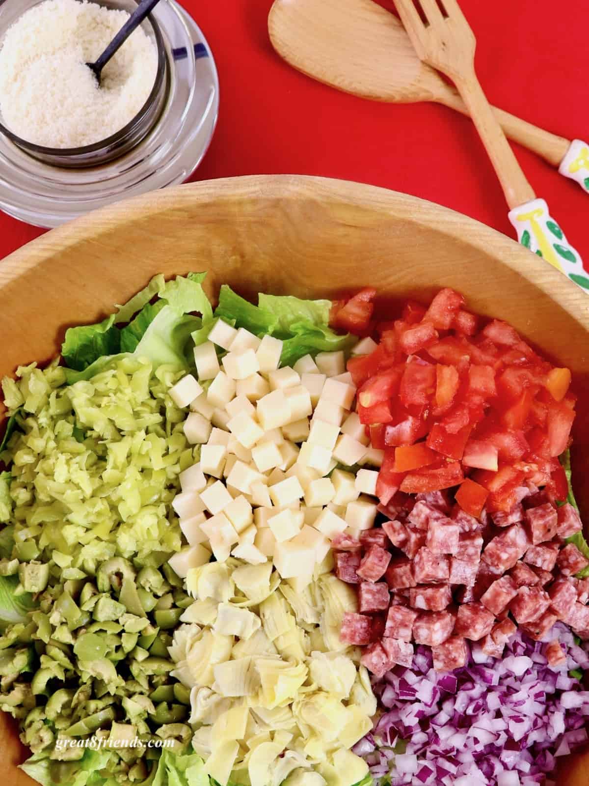 Wooden Salad Bowl with chopped tomatoes, salami, red onion, cheese, artichokes hearts, olives, and pepperoncini with salad servers and grated cheese.