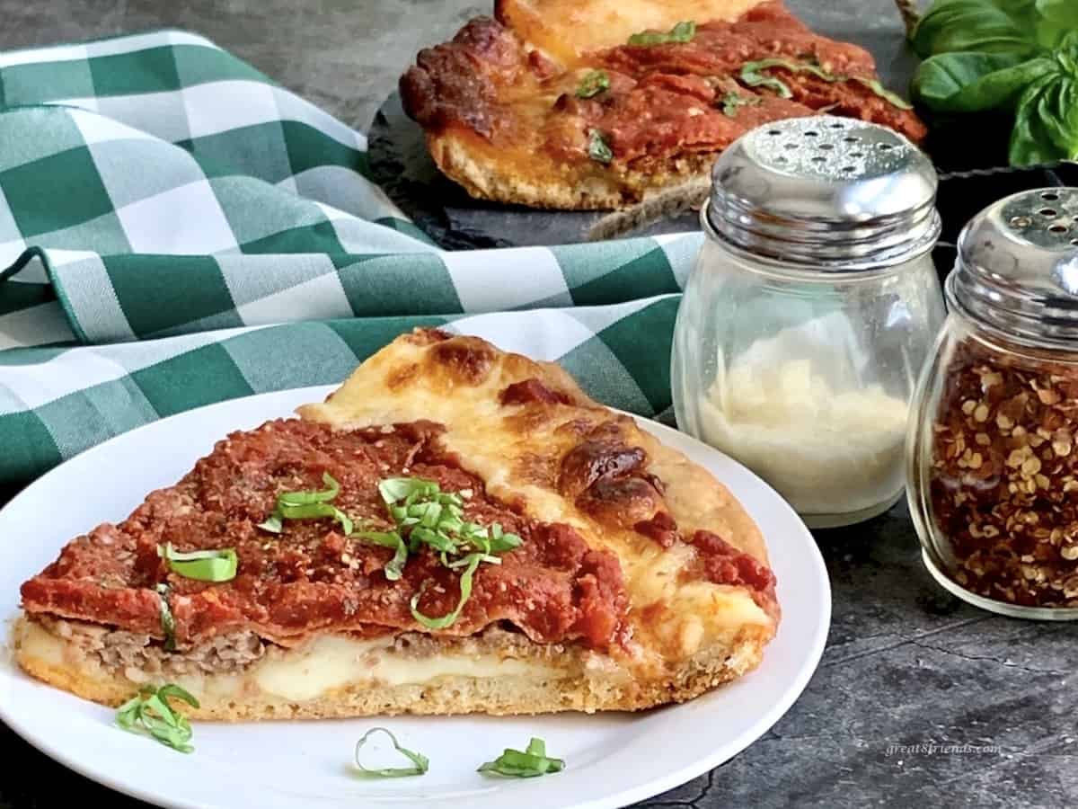 Thick chicago style pizza slice in the forefront and background with a green and white checked napkin on the table with parmesan and red pepper flakes on the side.