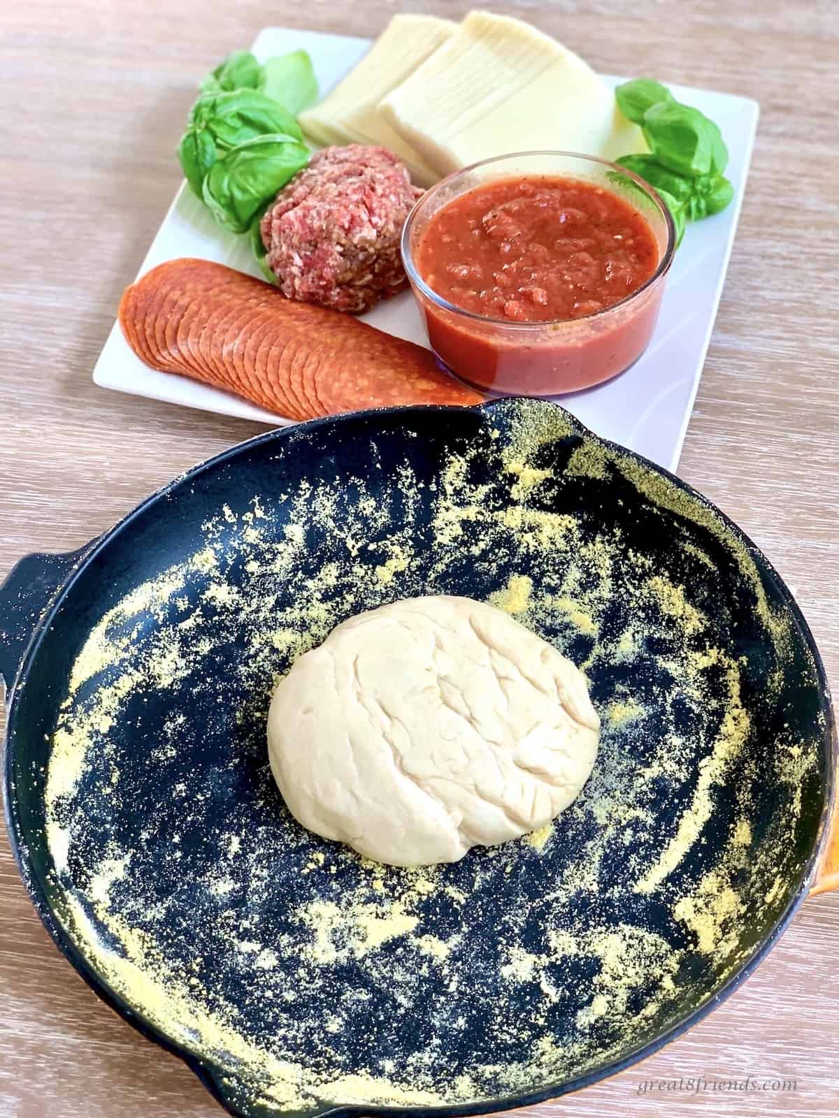 All ingredients for a Chicago pizza made in a cast iron skillet covered in corn meal. Ingredients on a white plate are sliced mozzarella, tomato sauce, sausage, pepperoni and basil leaves.