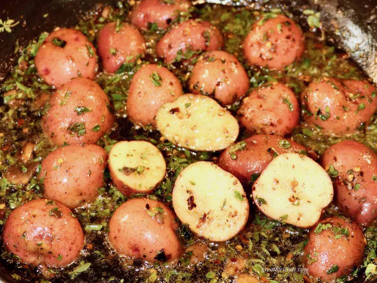 Halved red skinned potatoes and herbs browning in pan.