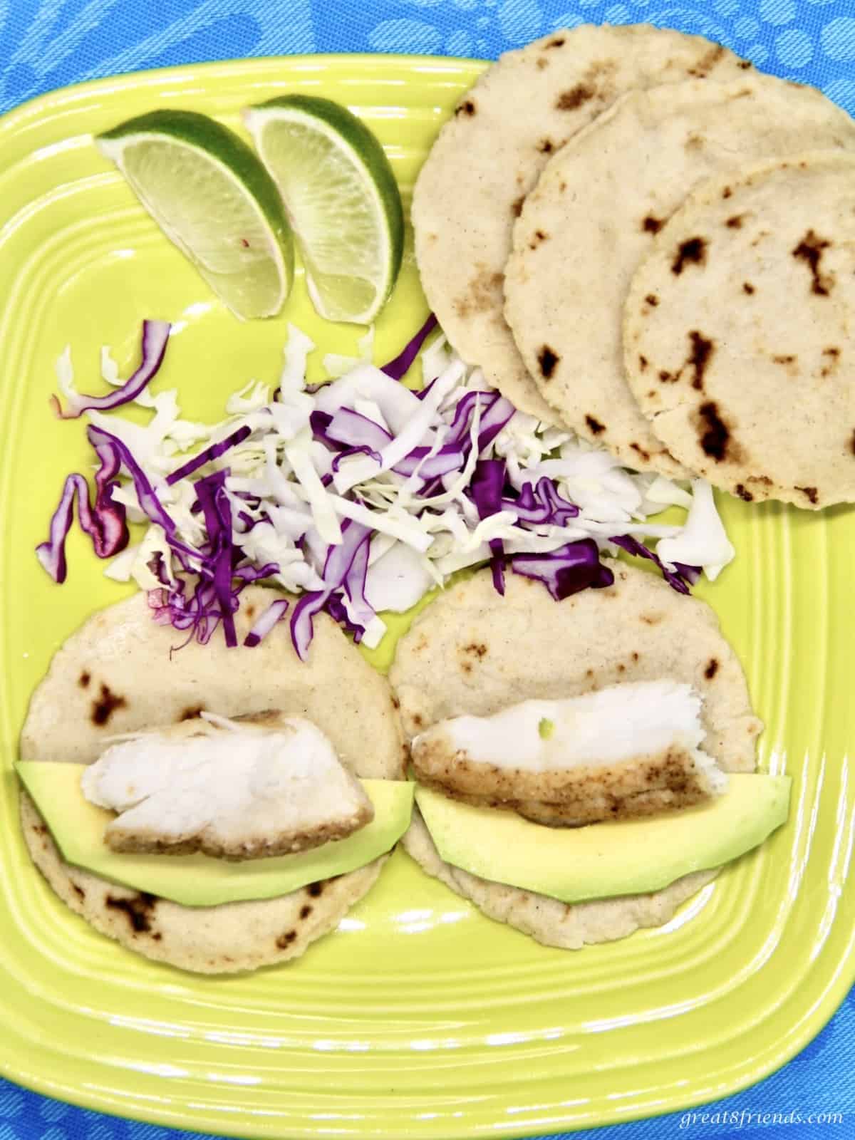 A bright yellow plate with two fish tacos with avocado, three corn tortillas on the side also with cole slaw and two lime slices.