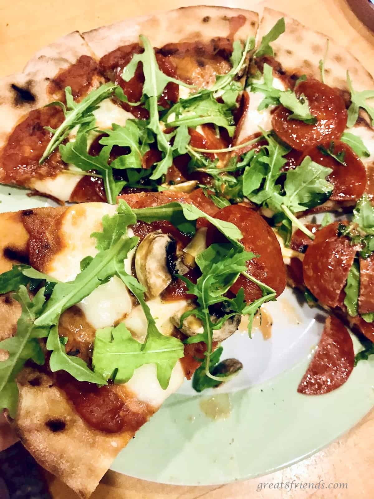 Upclose photo of a pizza cooked on the grill topped with sauce, cheese, pepperoni, and arugula.
