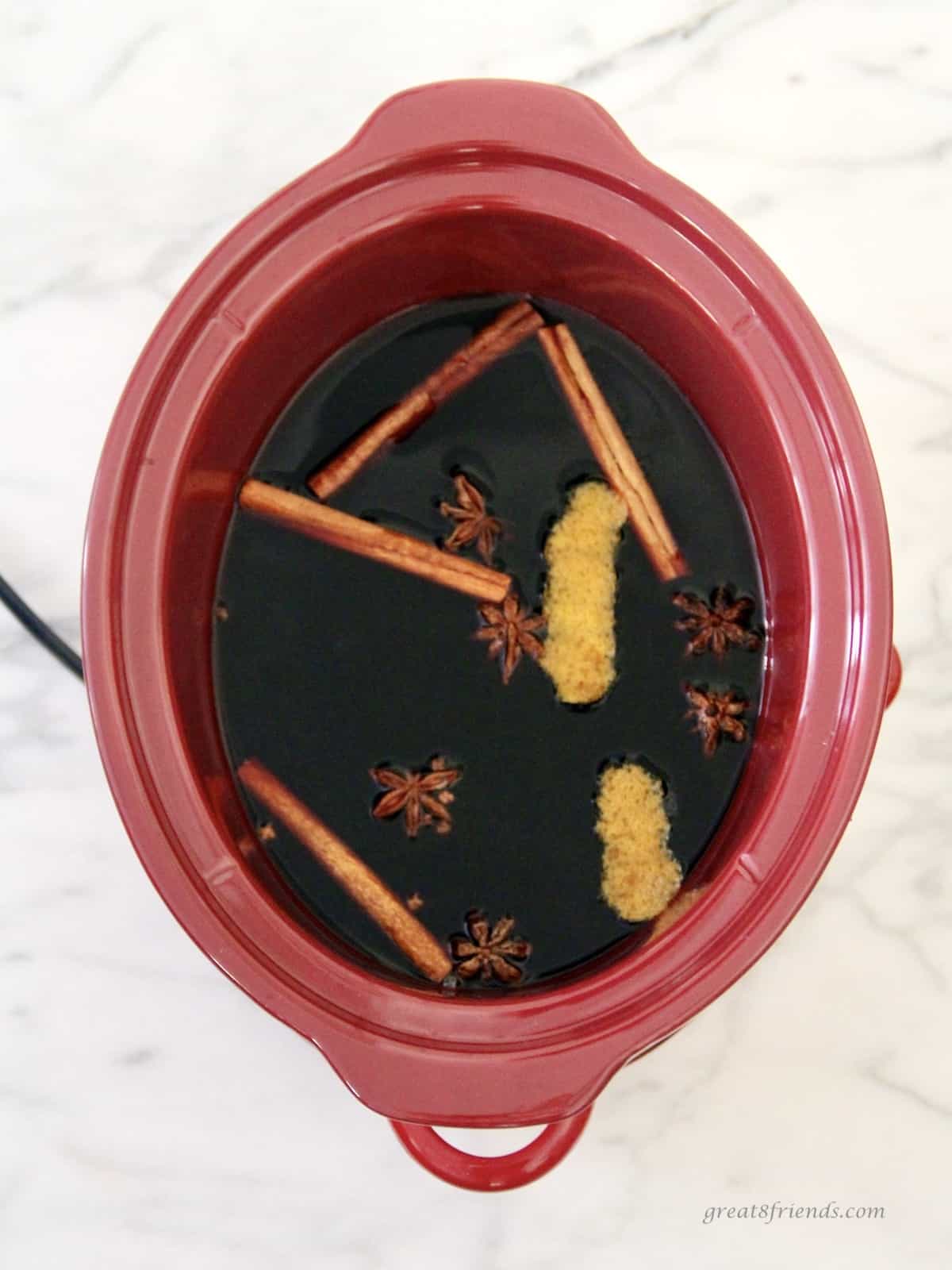 Overhead view of a red slow cooker with red wine, cinnamon sticks, orange zest, whole cloves and star anise.