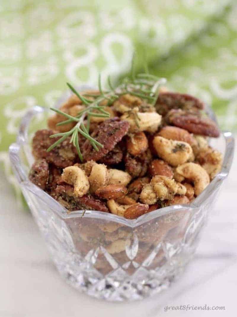 Rosemary spice nuts in a glass bowl garnished with a sprig of rosemary.