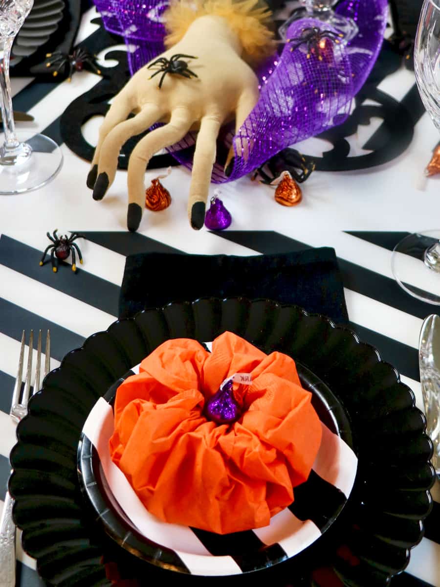 Halloween table setting with striped mat, black plate and a paper napkin pumpkin. Creepy hand with spiders in the background.