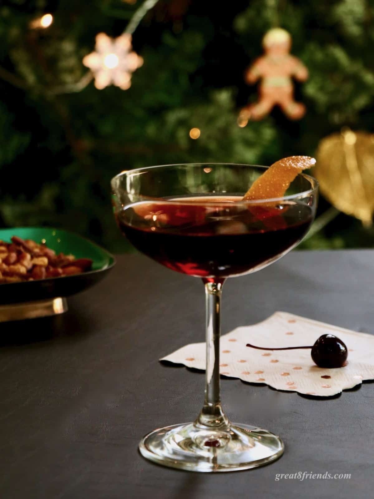 A cherry Manhattan in a coupe glass garnished with orange peel with a cherry on the table and nuts in background.