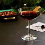 A cherry Manhattan in a coupe glass garnished with orange peel with a cherry on the table and nuts in background.