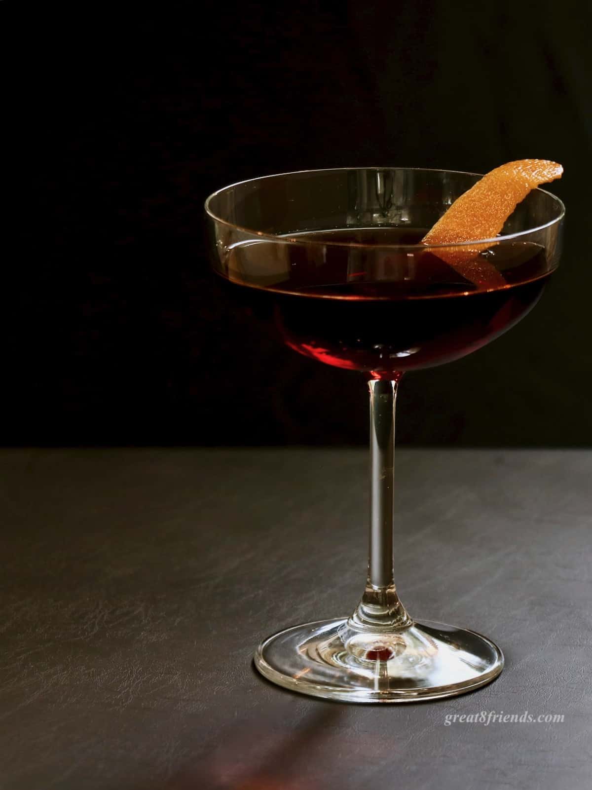 A cherry Manhattan in a coupe glass garnished with orange peel.