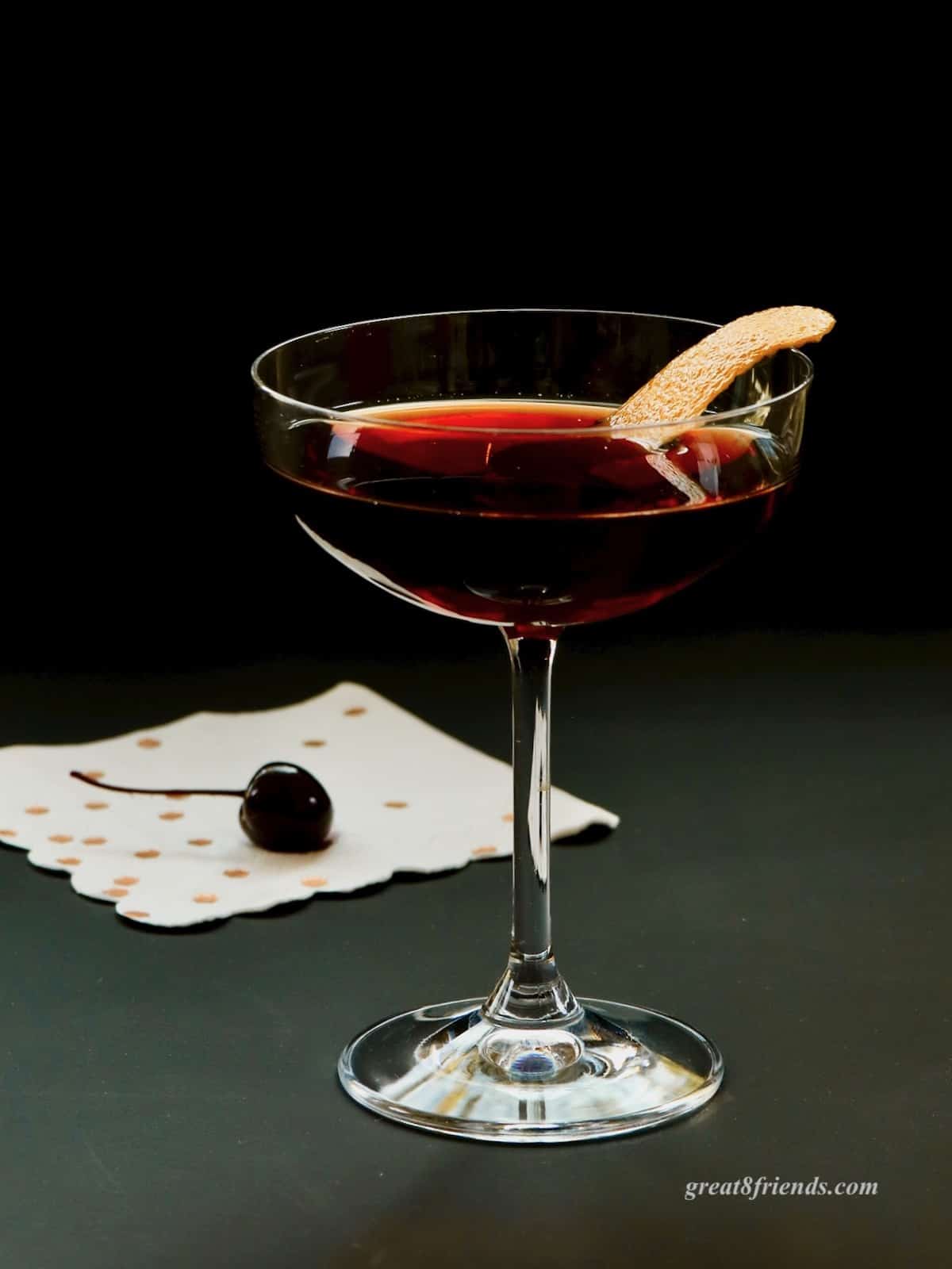 A cherry Manhattan in a coupe glass garnished with orange peel with a cherry on a napkin.