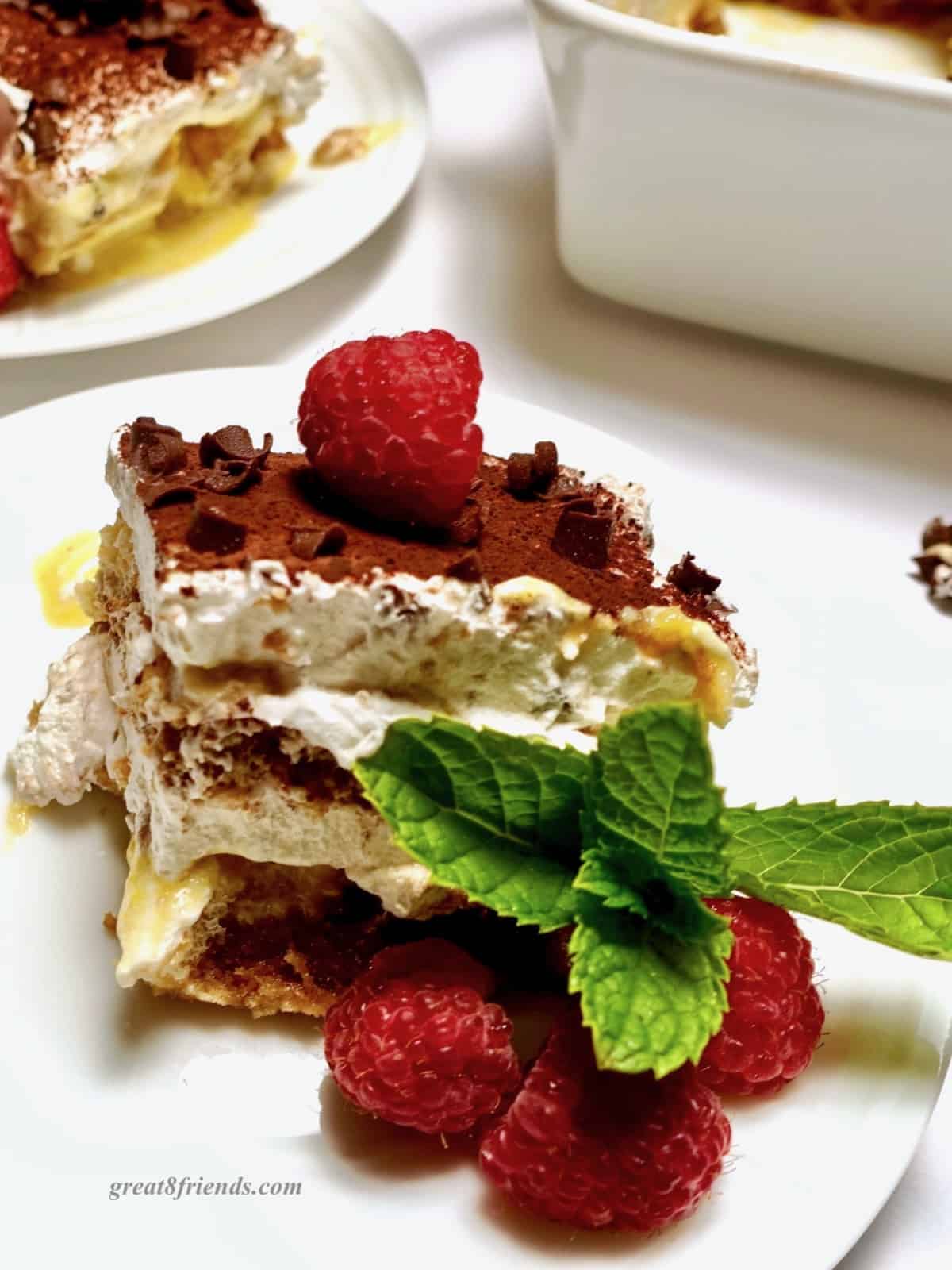 A piece of tiramisu on a plate garnished with raspberries and mint sprigs.