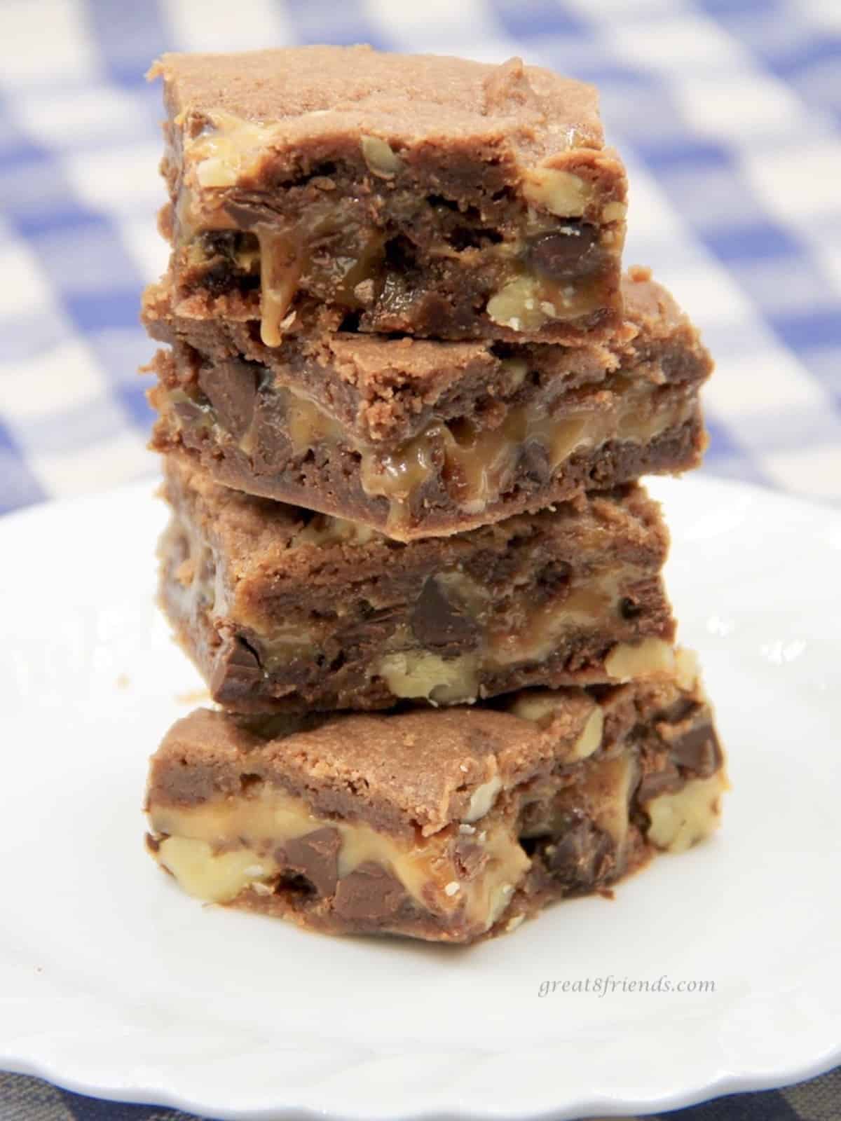 Four brownies made with caramel stacked on top of each other on a white plate on a blue and white checked tablecloth.