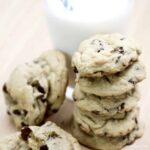Several chocolate chip cookies stacked on each other in front of a glass of milk with a blue straw in it.