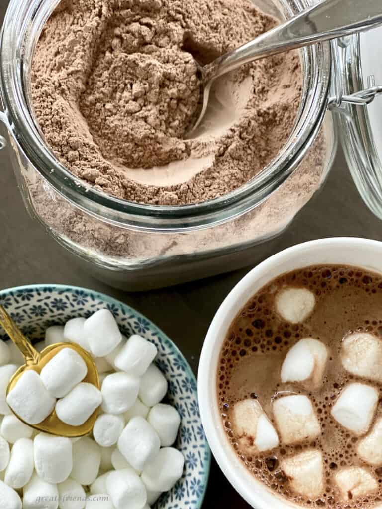 Overhead view of powdered cocoa mix, a cup of hot cocoa with marshmallow on top, and a bowl of mini marshmallows.