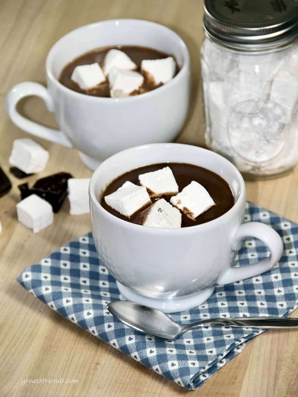 Two white mugs with homemade hot cocoa each with four small square homemade marshmallows sitting on a blue cloth napkin with a jar of marshmallows in the background.