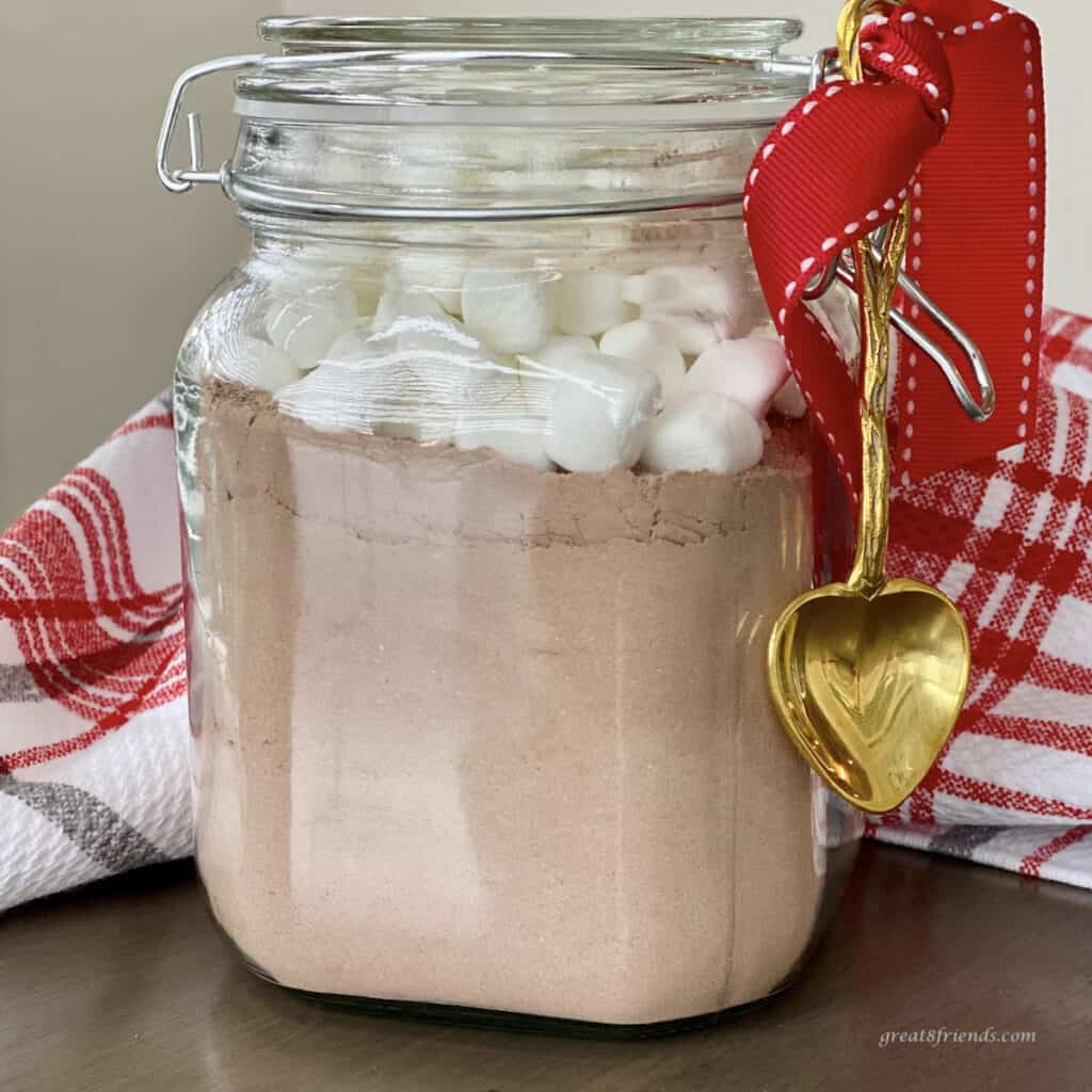 Hot cocoa mix in a jar with marshmallow on top wrapped as a gift with a red bow with a gold spoon hanging on the side.