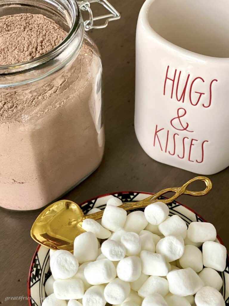 A jar of homemade cocoa mix next to a mug saying "hugs & kisses" and a bowl of mini marshmallows with a gold colored spoon.