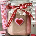 Powdered cocoa mix in a mason like jar with a ribbon tied around it and heart paper cups in the background.