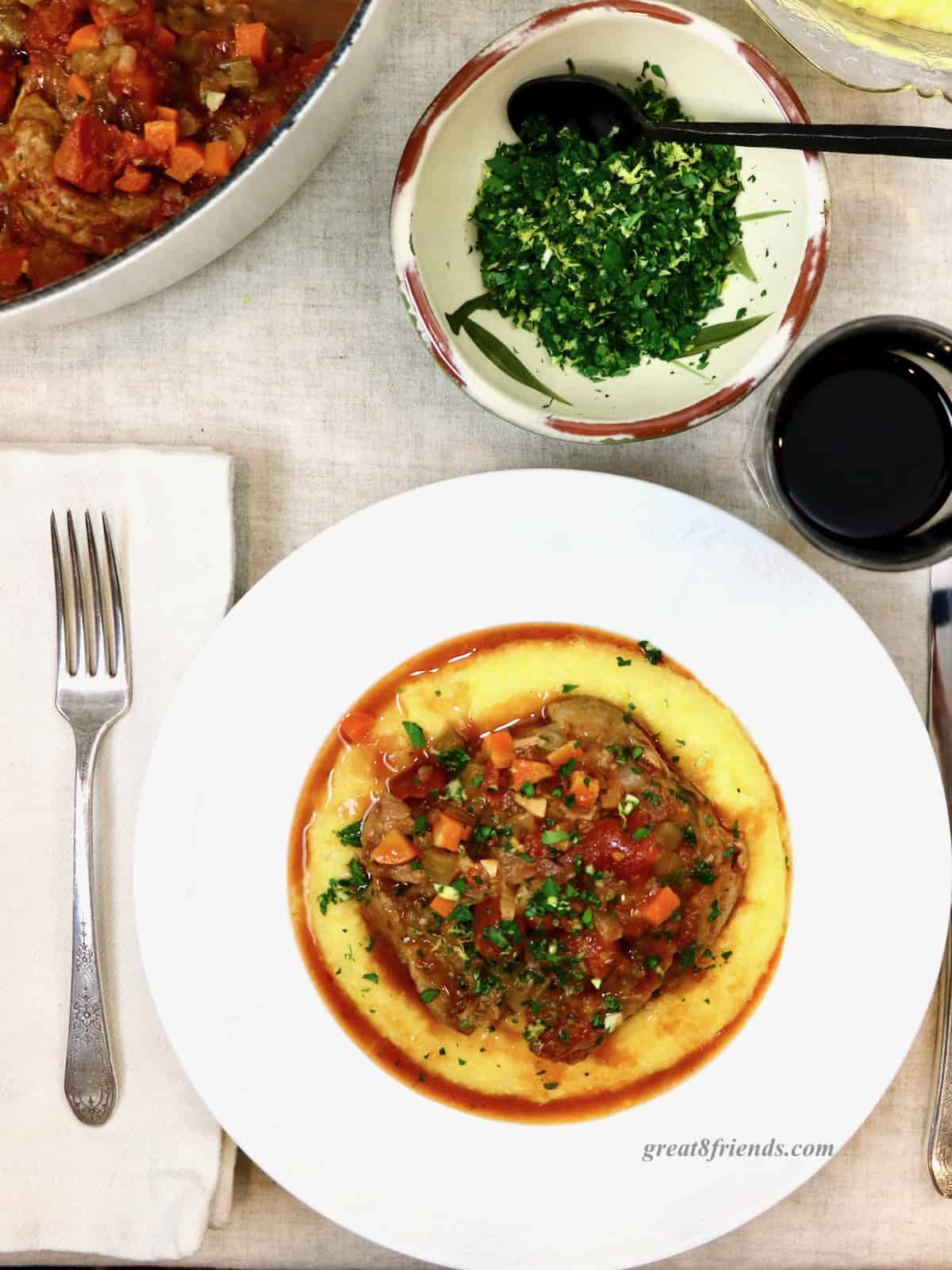 A place setting with osso buco on a bed of polenta in a white dish topped with gremolata. With a dish of gremolata above the plate.