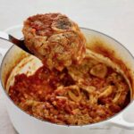 A close up of a piece of osso buco being lifted from the white pot with the rest of the osso buco.