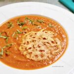 Tomato soup in a white rimmed bowl topped with shredded basil and a parmesan crisp.