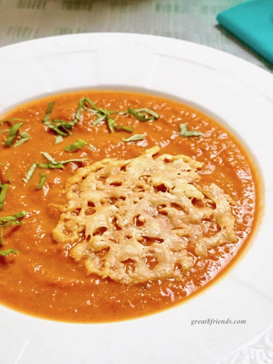 Tomato soup in a white rimmed bowl topped with shredded basil and a parmesan crisp.