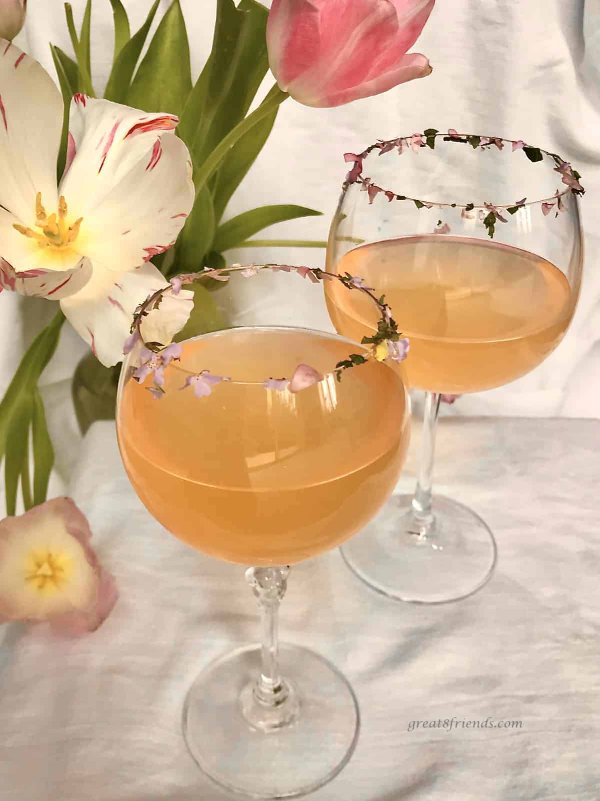 Two glasses of pink lemonade with edible flower bits on the rim of the glasses and a tulip flower on the table for decoration.