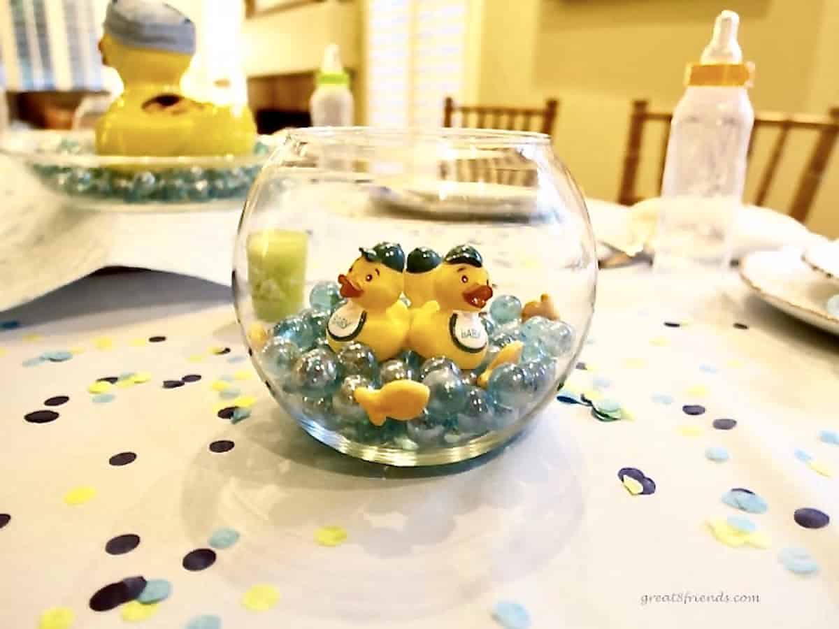 On a decorated table with confetti a glass fish bowl with blue glass marbles and rubber ducks inside.