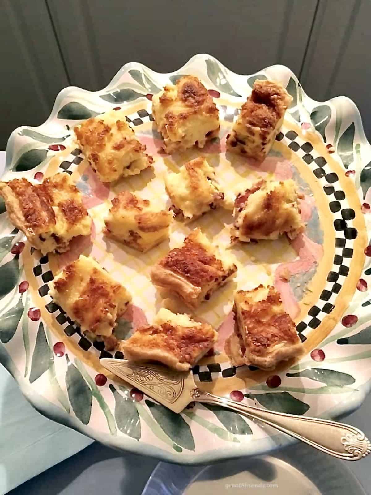 Quiche cut into small squares and served on a colorful round platter with a small silver spatula.