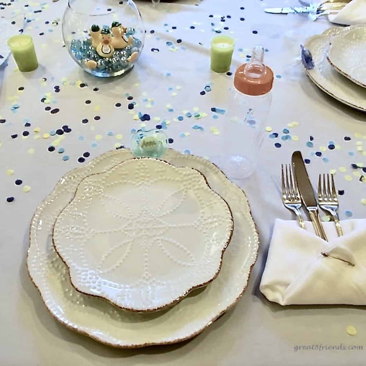 Table setting with confetti on a white table cloth, a baby bottle and a fish bowl with glass marbles and small rubber ducks.