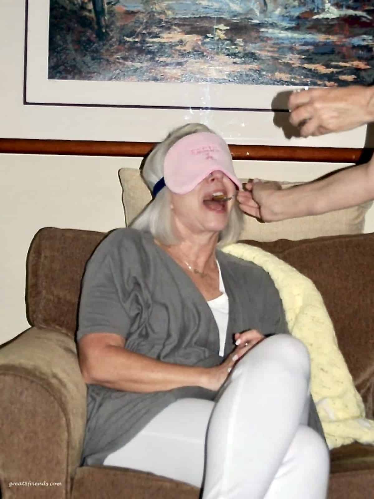 A woman with a pink blindfold on her eyes while someone is feeding her with a spoon.