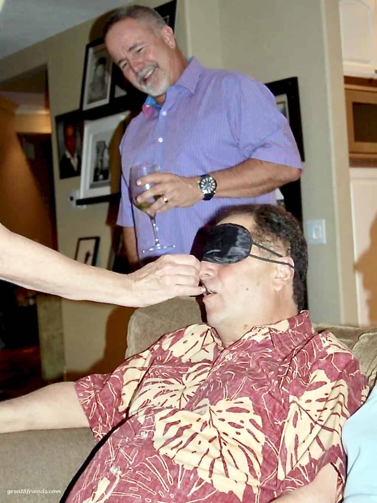 A man with a blindfold on his eyes while someone is feeding him and another man in the background watching with a smile.