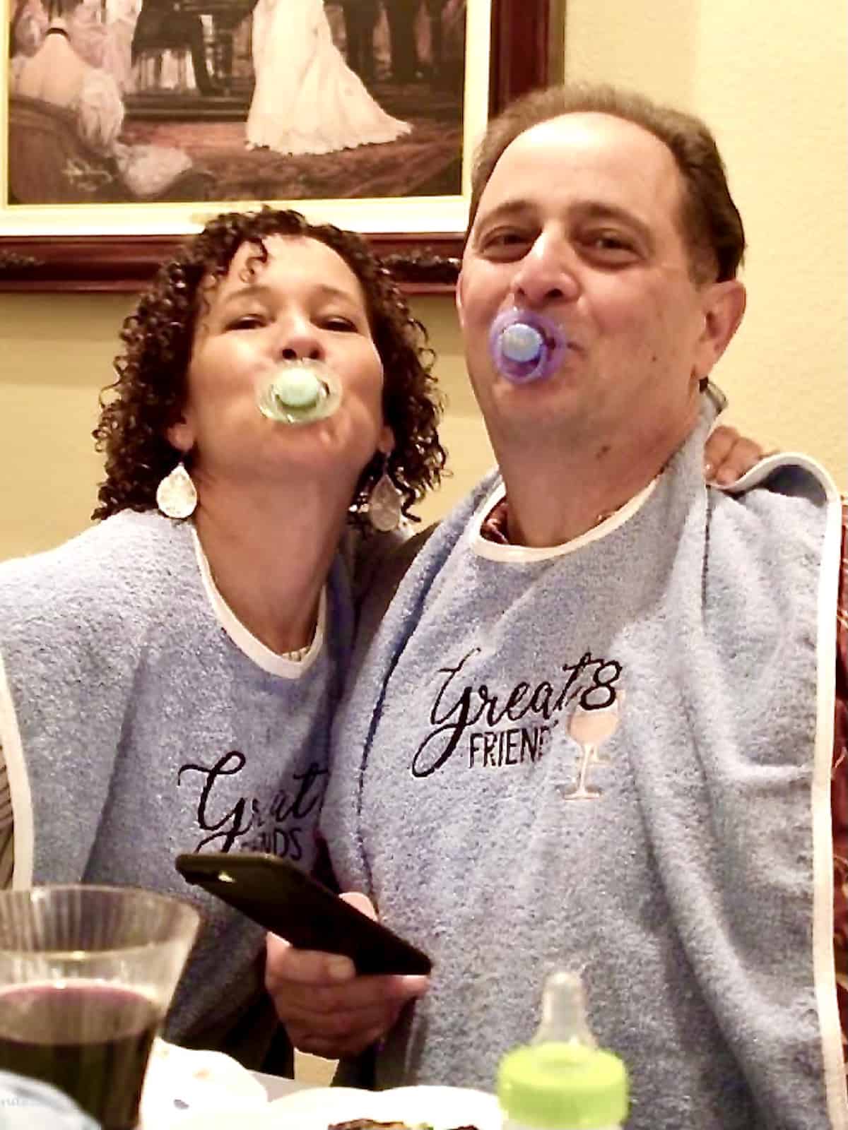 Two adults posing for the camera with a pacifier in their mouth and adult bibs on.