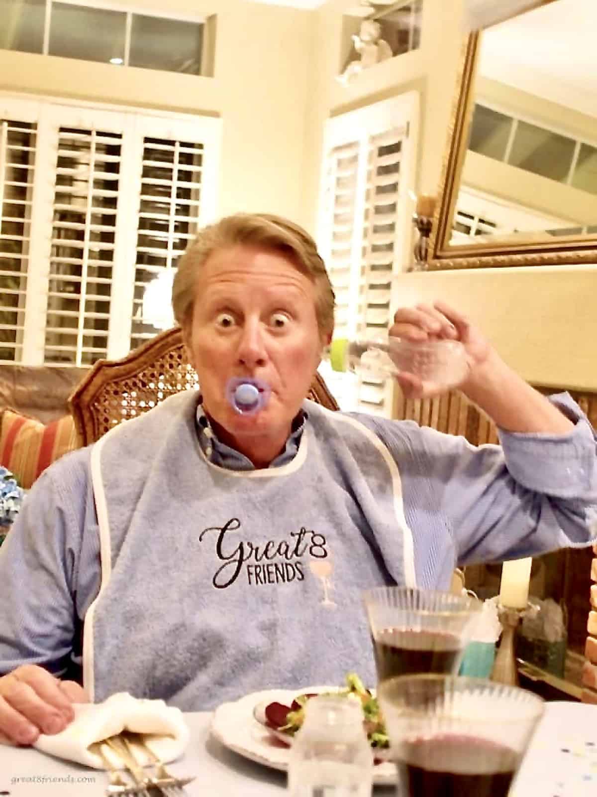 A man with a pacifier in his mouth, a baby bottle in his ear wearing an adult bib and sitting at a table with food and wine.