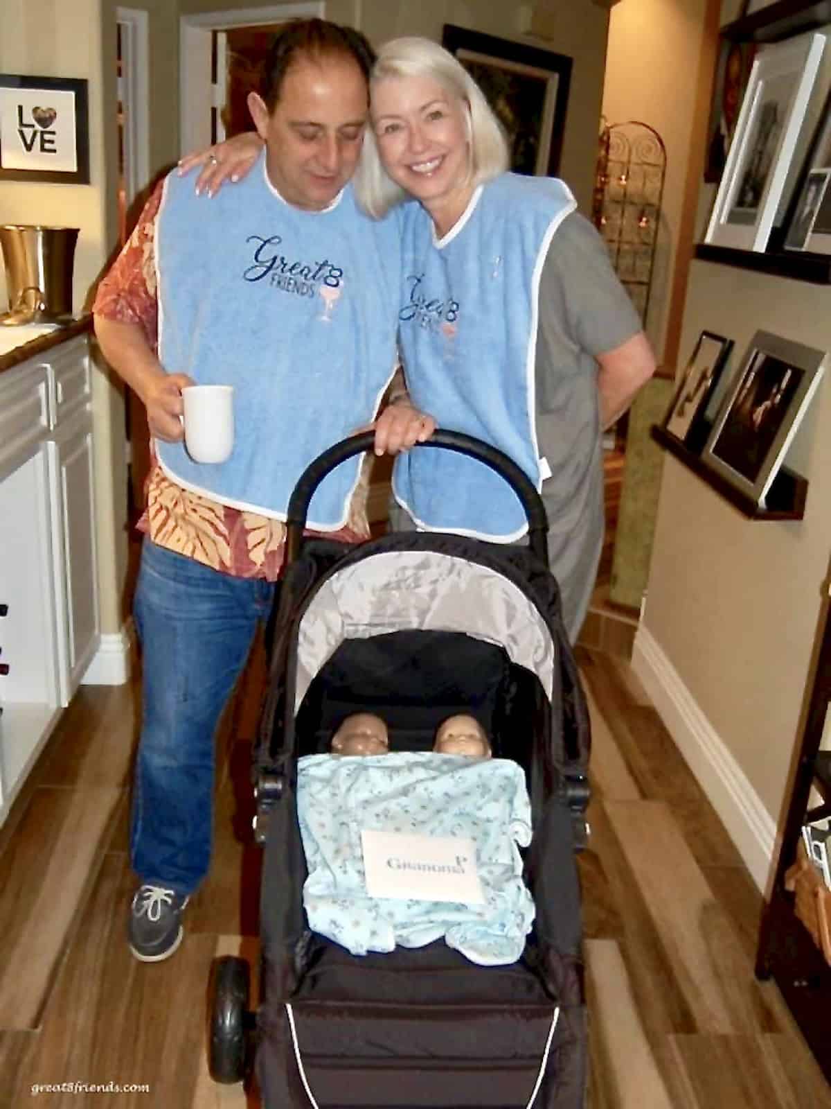 A man and a woman standing behind a black stroller with two baby dolls in it covered with a baby blanket.