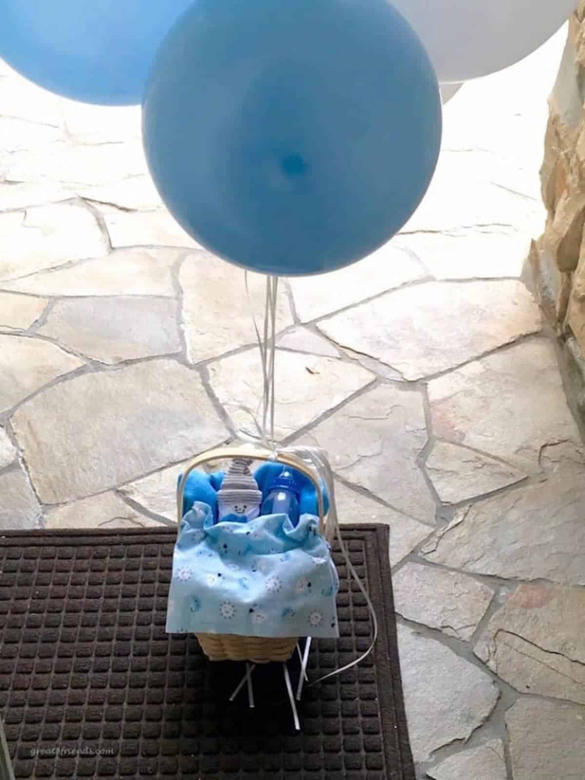 A basket with a baby bottle and a blue baby blanket on a porch with a blue balloon tied to the handle.