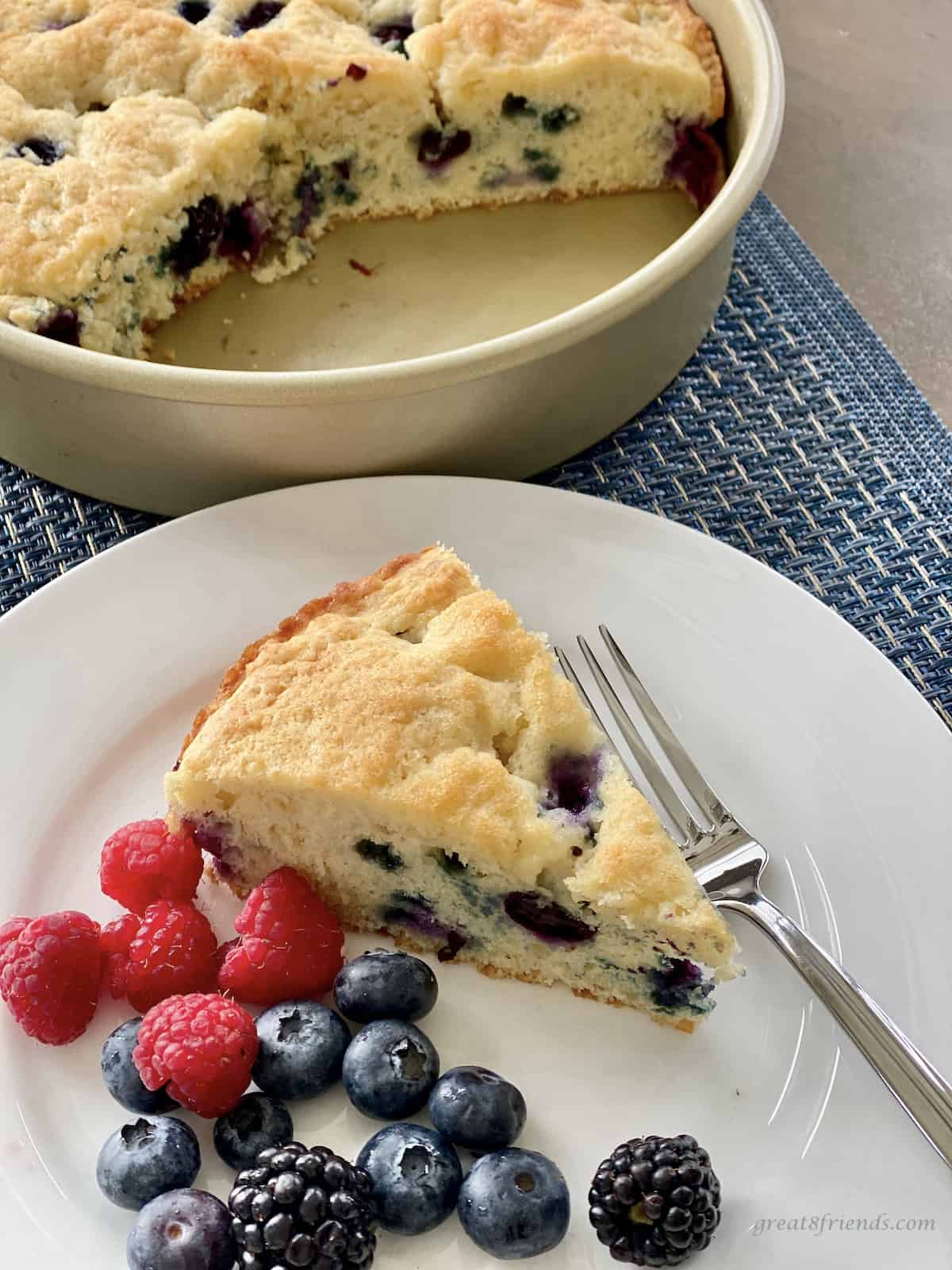 A piece of blueberry coffee cake on a plate with fresh blueberries and raspberries with a round cake pan with a piece of the coffee cake cut out of it.