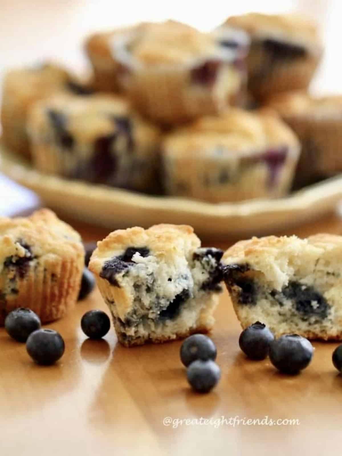 Blueberry muffins on the table in front of a stack of muffins on a plate with a few fresh blueberries on the table.