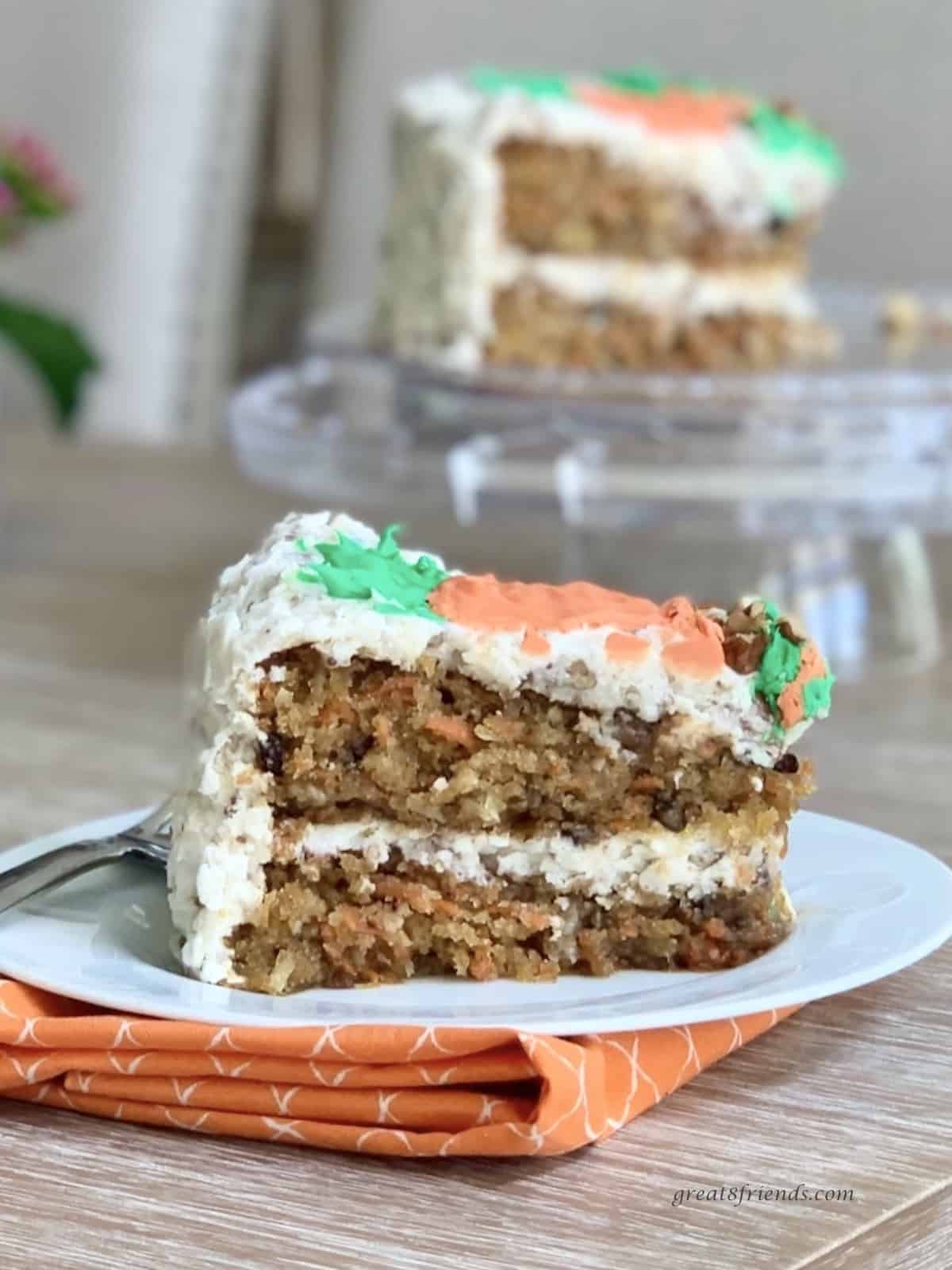 A piece of carrot cake on a white plate that is sitting on an orange cloth napkin with a carrot cake on a glass cake stand in the background.