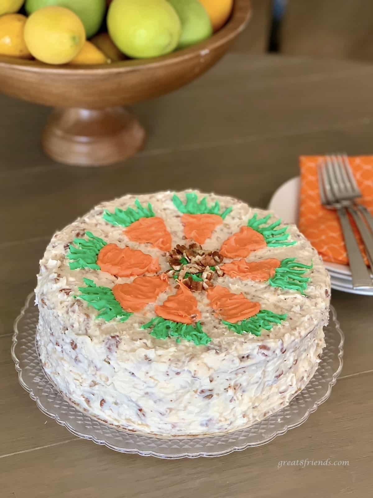 A carrot cake decorated with orange carrots topped with green icing carrot tops with a bowl of lemons in the background.