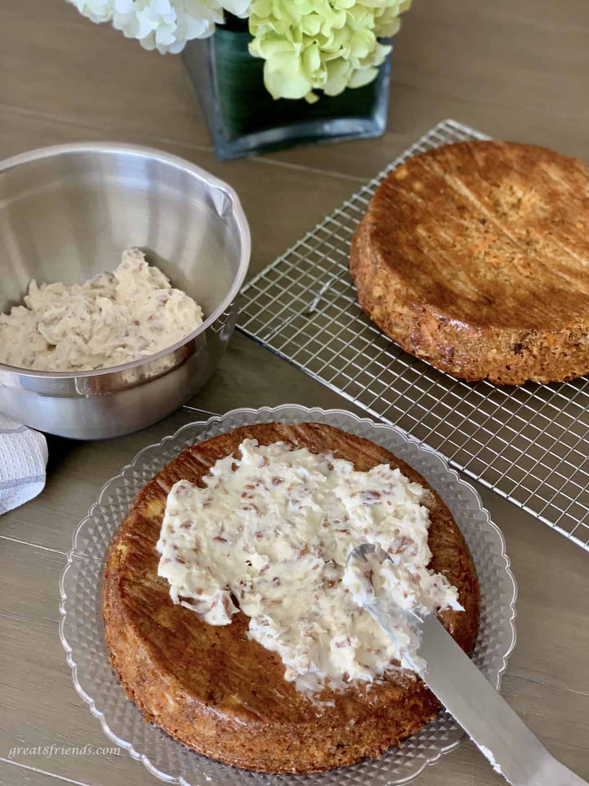 Two round baked cakes with one being frosted with a bowl of icing on the side.