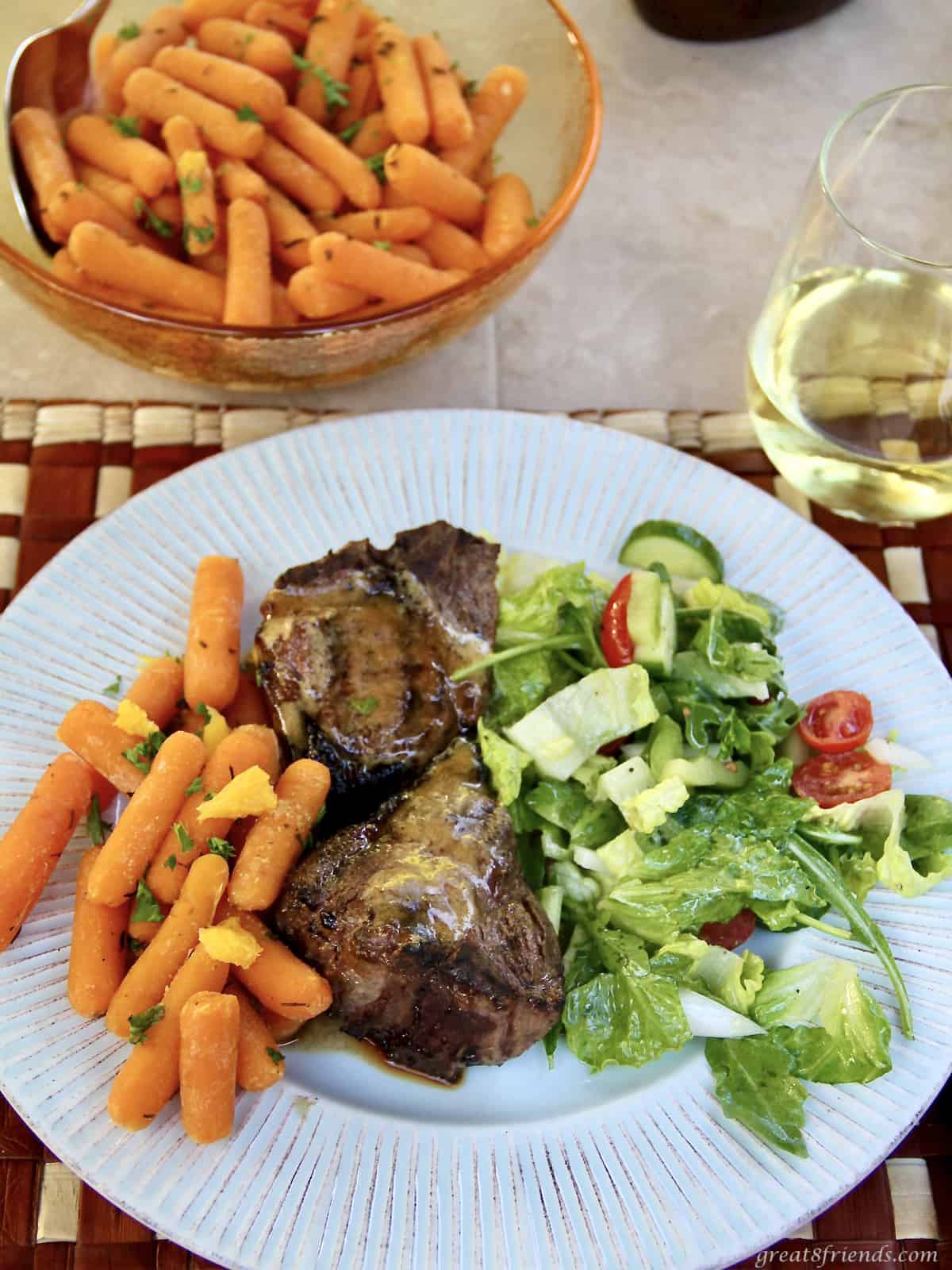 A plate with carrots, lamb chops and a green salad with a bowl of carrots and a glass of wine on the side.