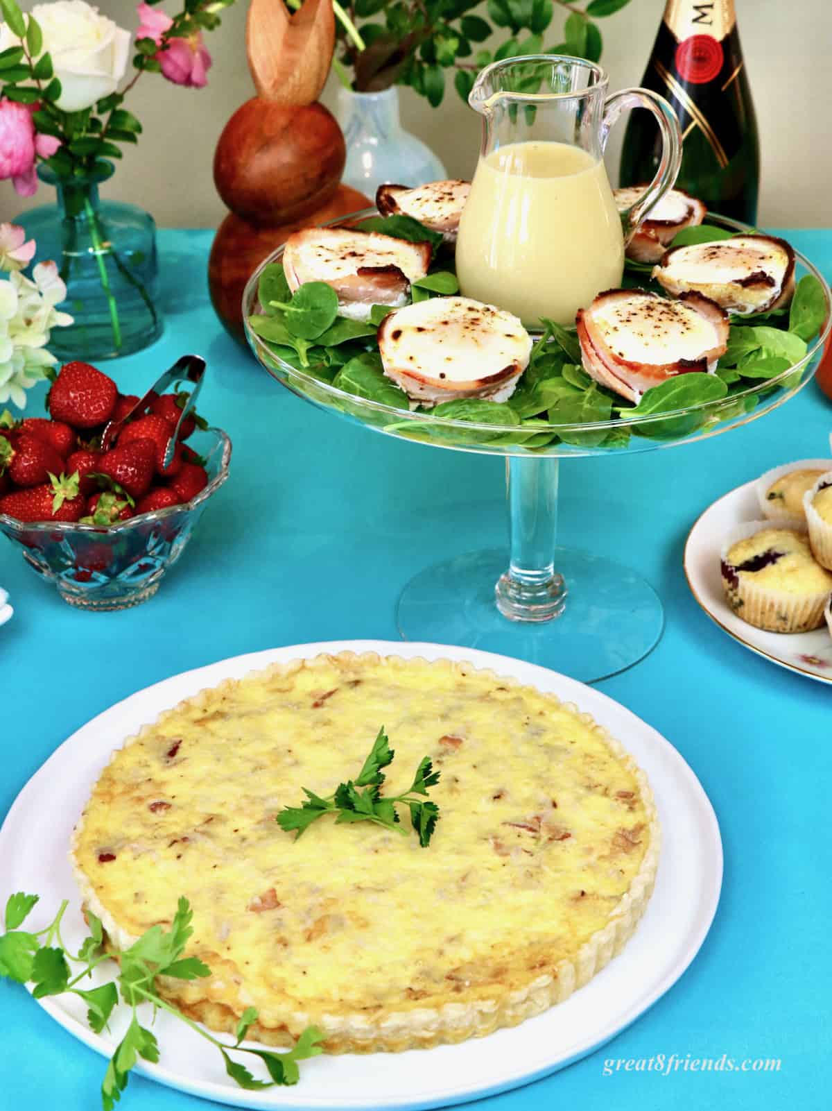 A whole quiche and a pedestal tray with eggs and a pitcher of hollandaise sauce on a robin's egg blue tablecloth.