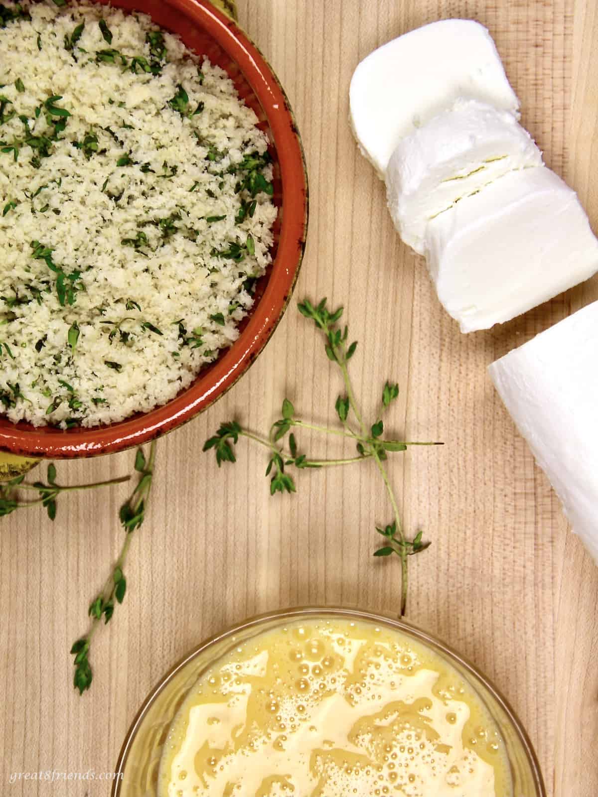 Bread crumbs in a red bowl and beaten egg in a glass bowl and cut goat cheese rounds on a wood board.